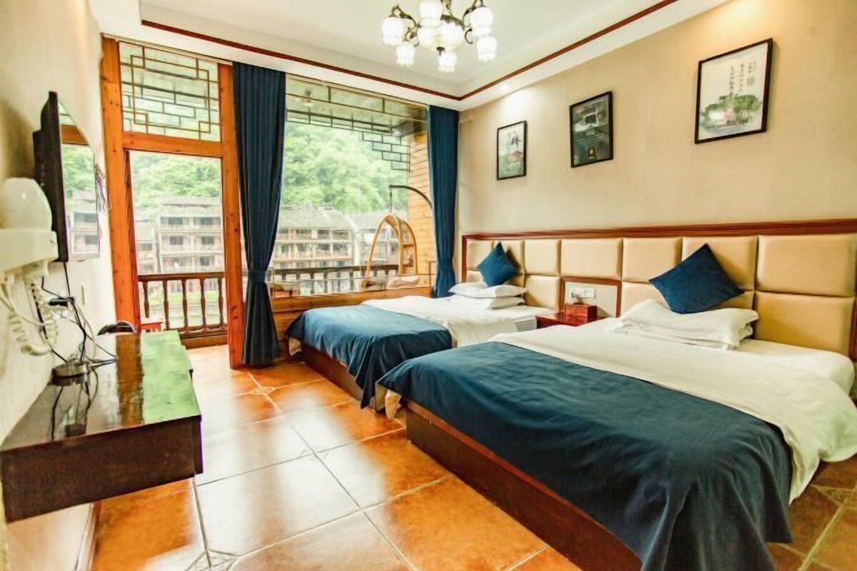 A hotel room with two beds, a desk, a chair, and a balcony with a view of buildings.