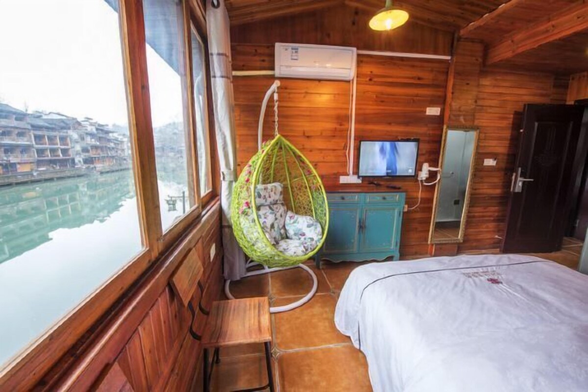 A wooden cabin room with a bed, a hanging chair, a TV, and a view of a river and buildings.