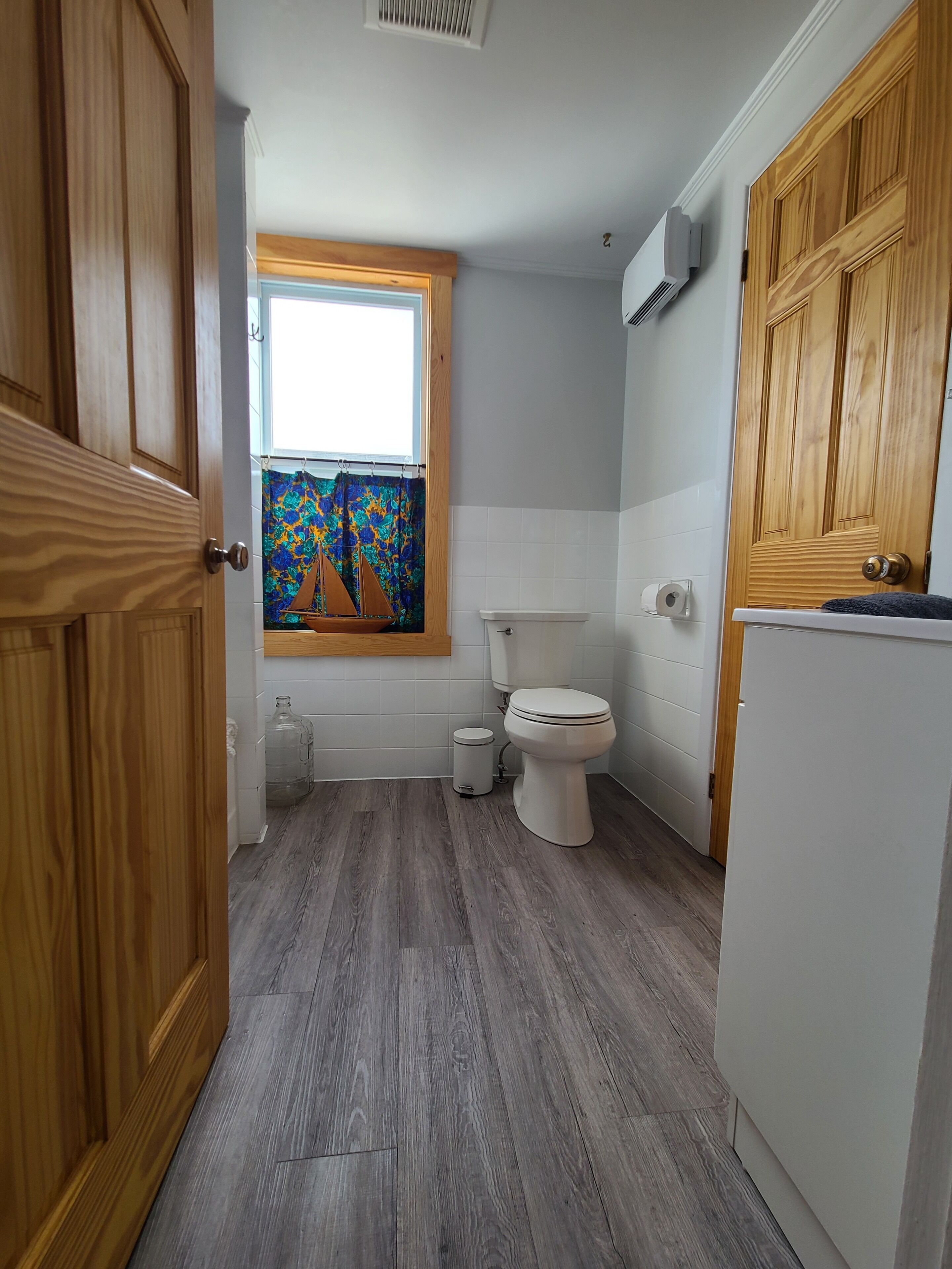 Une salle de bain avec des toilettes, une fenêtre ornée d’un rideau coloré, un grand distributeur d’eau et des armoires en bois.