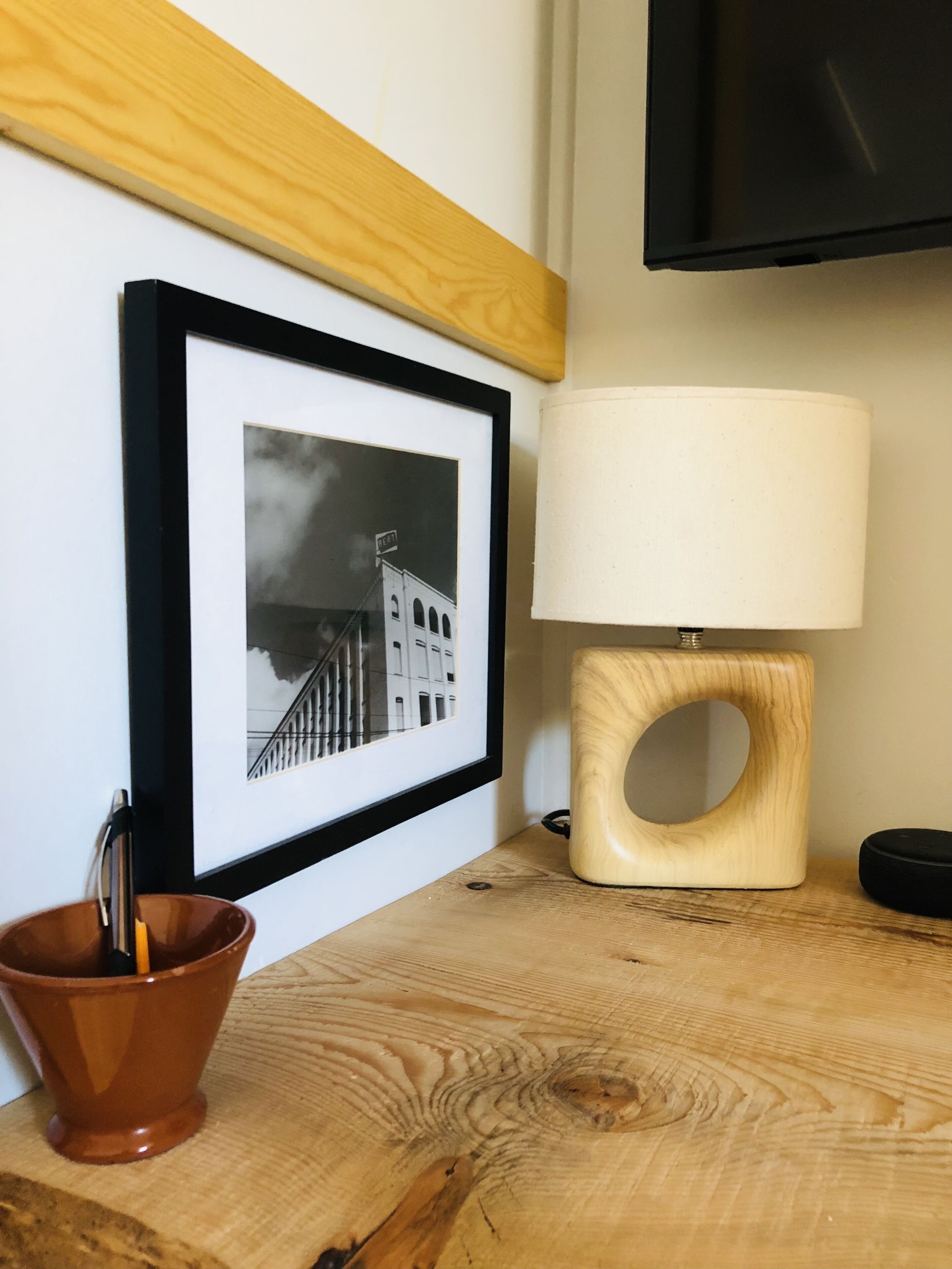Een lijst met een zwart-witfoto aan de muur, een houten tafel met een lamp en een bruine keramische mok.