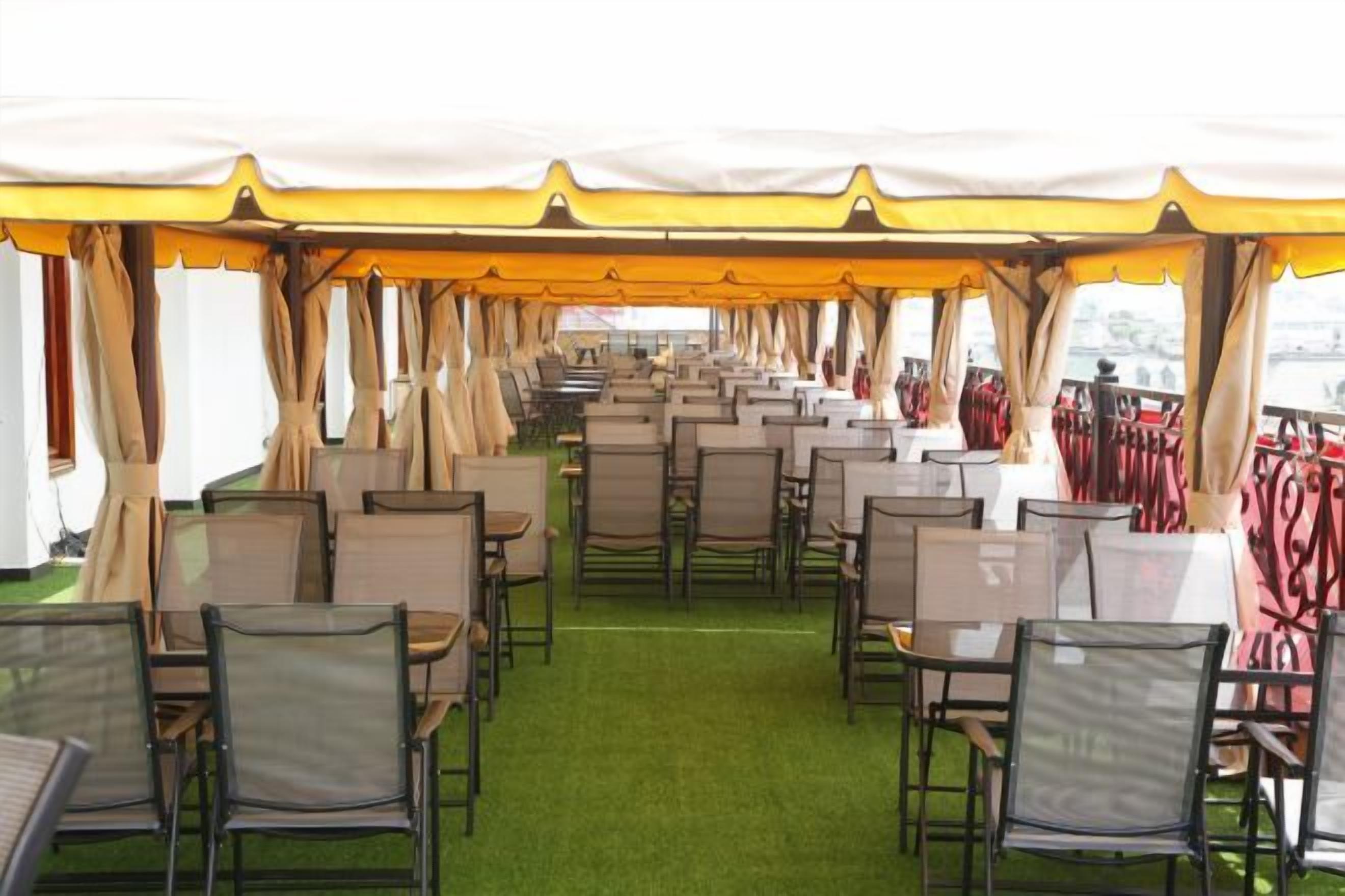 A tented dining area with rows of tables and chairs, green carpet flooring, and a view of the water outside.