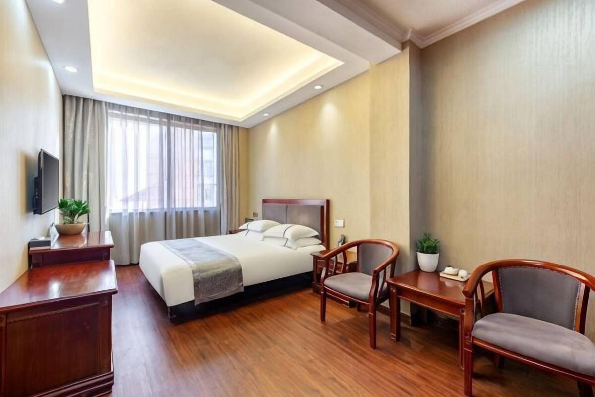 Une chambre d’hôtel avec un lit, deux chaises, un bureau, une télévision et une fenêtre avec des rideaux.