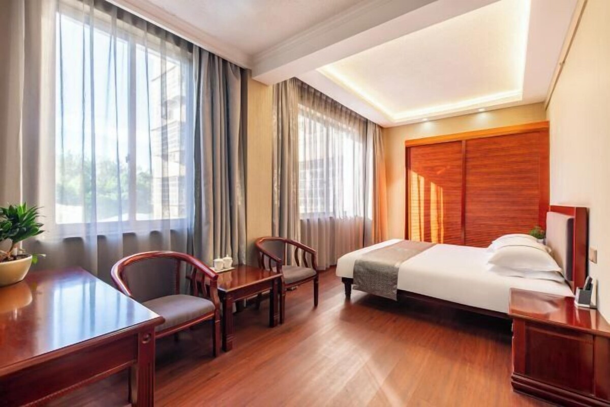Une chambre d’hôtel avec un lit, un bureau, deux chaises, une plante en pot et de grandes fenêtres avec des rideaux.