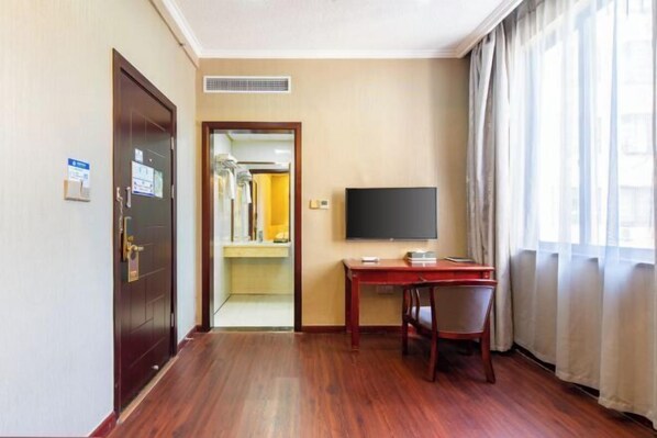 Une chambre d’hôtel avec un plancher en bois, un bureau avec une chaise, une télévision et une porte donnant sur un couloir où se trouvent un miroir et une étagère.