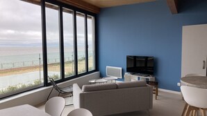Ein modernes Wohnzimmer mit großem Fenster zum Strandblick, einem Flachbildfernseher und einem Essbereich mit weißen Stühlen und einem Tisch.
