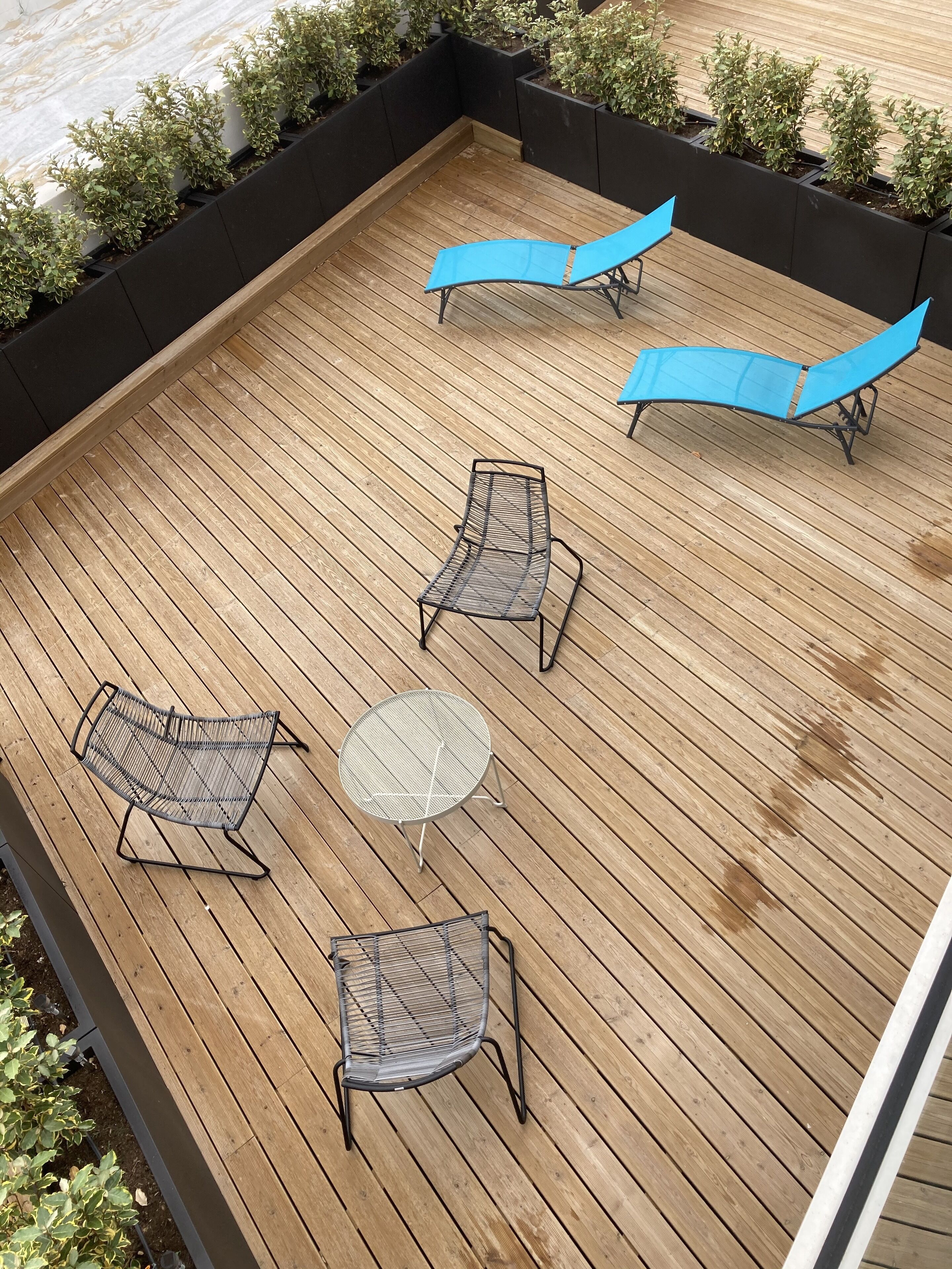 Une terrasse en bois avec quatre chaises en métal et une table ronde, entourée de jardinières noires dans lesquelles sont plantés des arbustes verts.