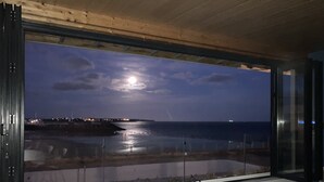 Ein modernes Wohnzimmer mit einem großen Fenster mit Blick auf einen Strand bei Nacht, das einen mondbeschienenen Meeresblick bietet.