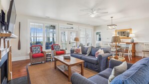 A spacious living room with a fireplace, large windows, and a ceiling fan.
