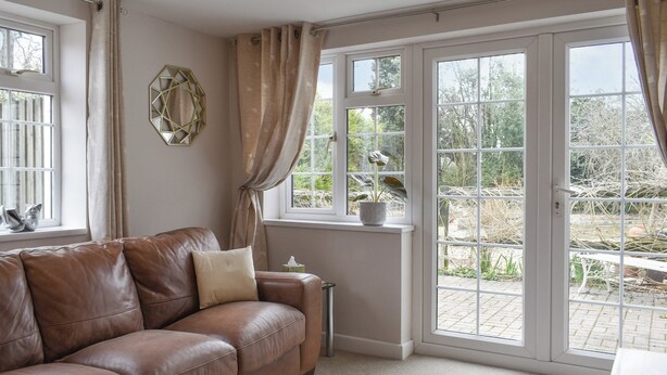 A room with a brown leather sofa, a small table, a potted plant, and a view of a garden through large windows. - 1 bedroom accommodation in Sway, near Lymington (Sway, near Lymington)