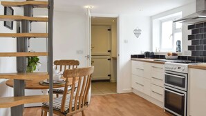 A modern kitchen with white cabinets, a stove, oven, and a dining area with a wooden chair and table. - 1 bedroom accommodation in Sway, near Lymington (Sway, near Lymington)