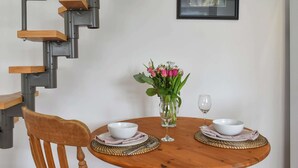 A dining area with a round wooden table set for two, a vase of pink flowers, and a framed picture on the wall. - 1 bedroom accommodation in Sway, near Lymington (Sway, near Lymington)