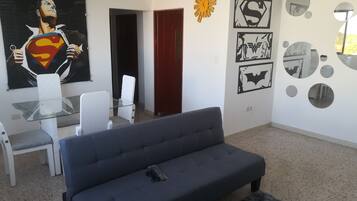 A modern living room with a grey sofa, a glass coffee table, white chairs, and a Superman poster. There are wall decorations including a sun-like shape and Batman-themed artwork.