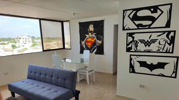 Quarto com sofá azul, mesa de vidro e cadeiras, uma janela ampla com vista para edifícios e paredes decoradas com logotipos de super-heróis.
