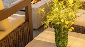 Un área junto a la piscina con una chimenea, mobiliario exterior y un florero con flores amarillas.