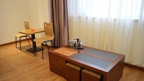 Una habitación de hotel con una mesa de comedor de madera y sillas, una pequeña kitchenette con hervidor y cafetera, y un ventanal con cortinas.