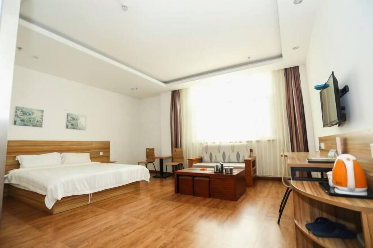 A hotel room with a wooden bed, a desk with a chair, a coffee table, a TV mounted on the wall, and a window with curtains.