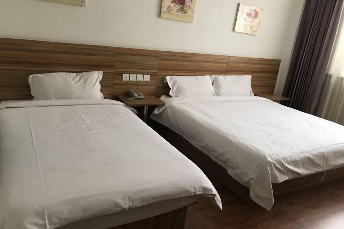 Two beds with wooden headboards, white bedding, a bedside table with a phone, and wall-mounted light switches.