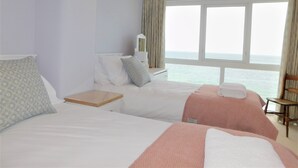 Una camera d'albergo con due letti, un comodino, una sedia, una lampada e vista sul mare.