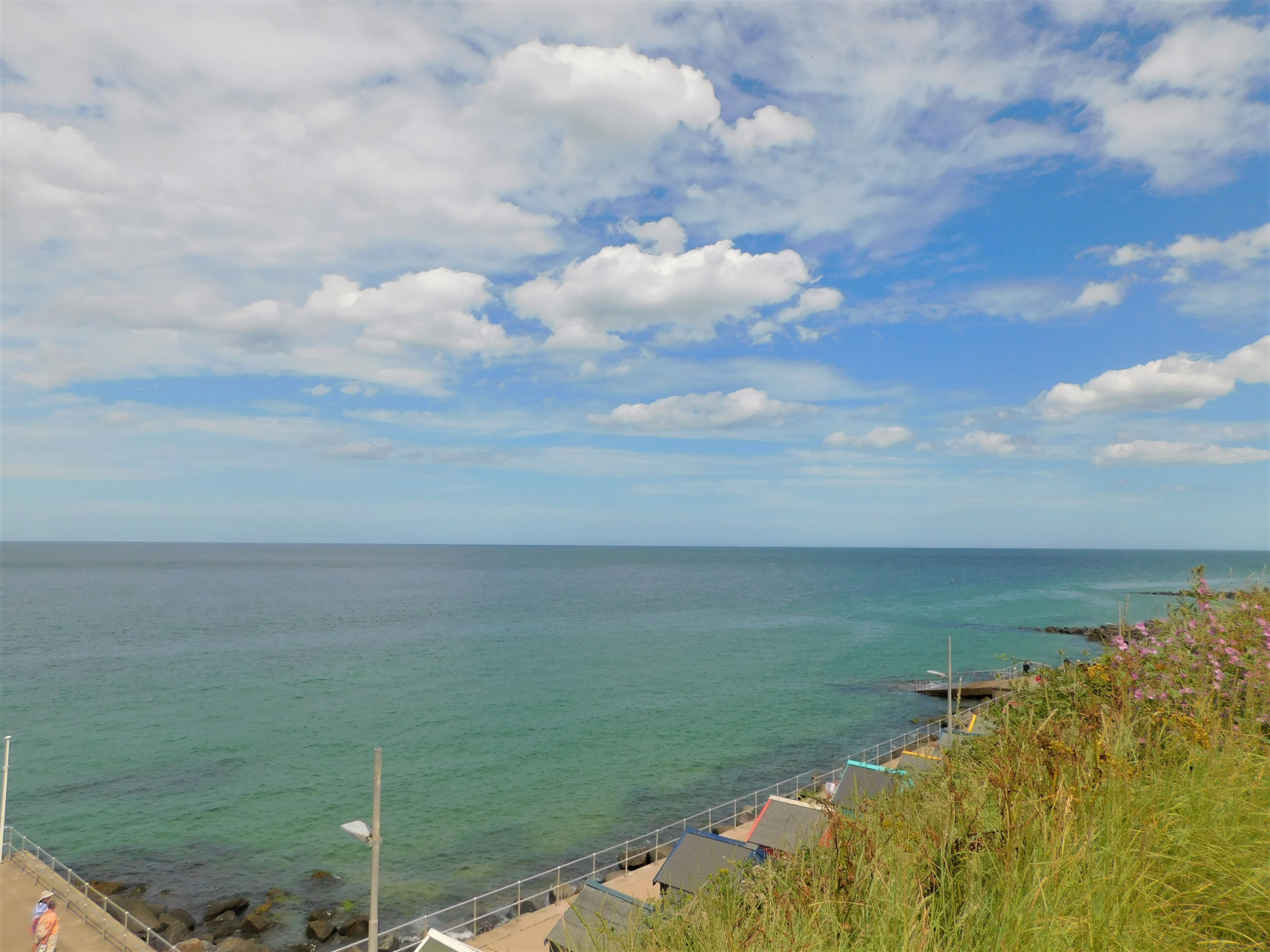 A coastal view with a clear sky, calm sea, and a rocky shoreline with a walkway.
