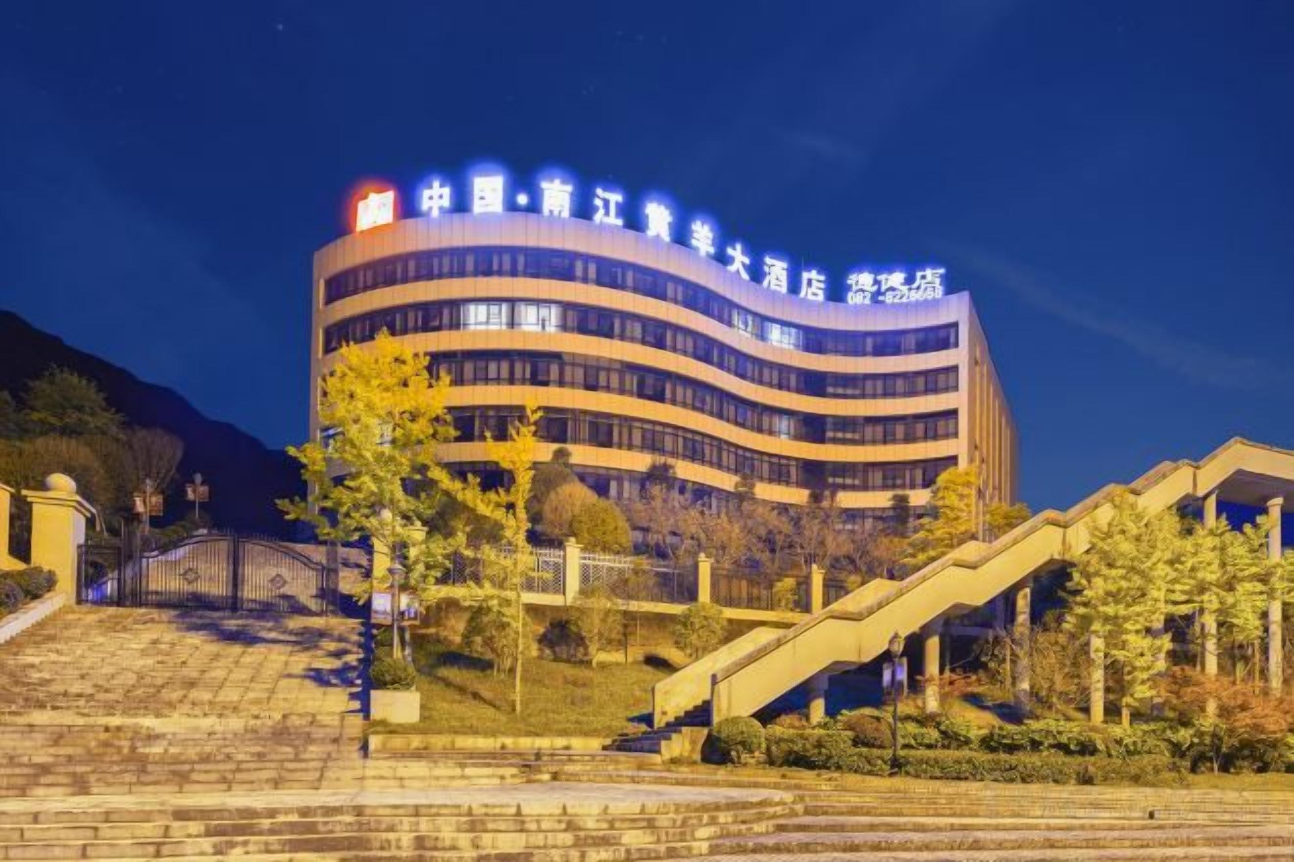 A multi-story hotel with a curved facade, a gated entrance, and a landscaped area with stairs and trees.