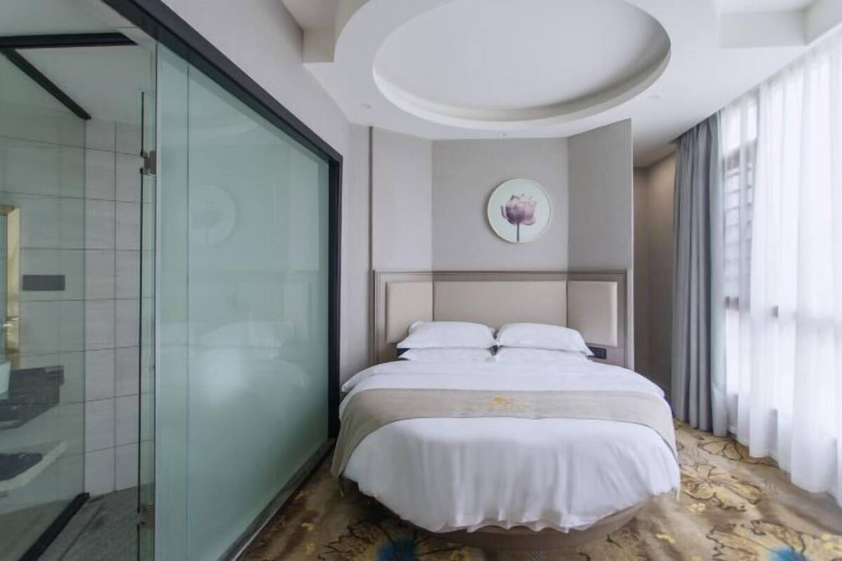 A hotel room with a bed, a glass shower enclosure, and a large window with curtains.