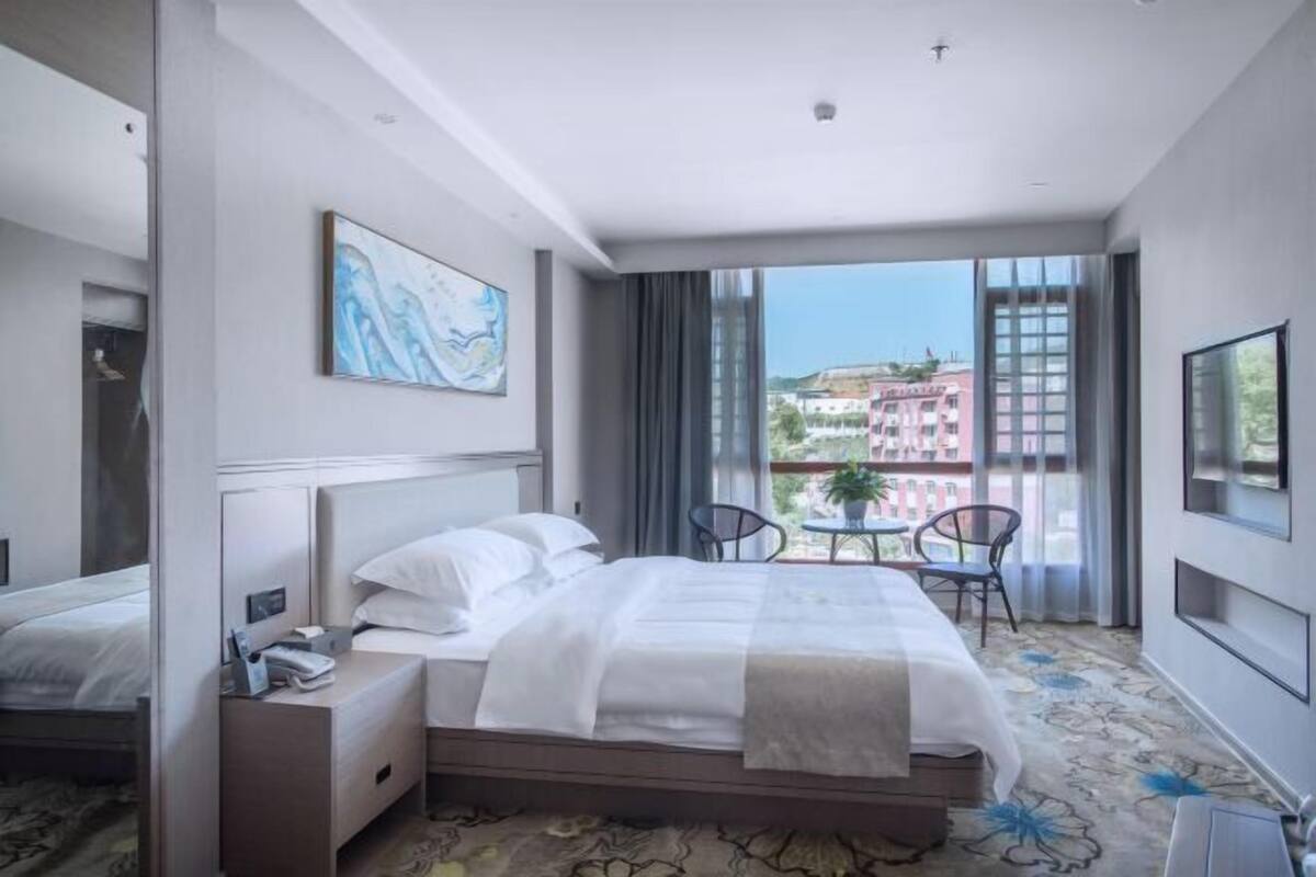 A hotel room with a bed, a nightstand, a chair, a table, and a large window with a view of buildings.