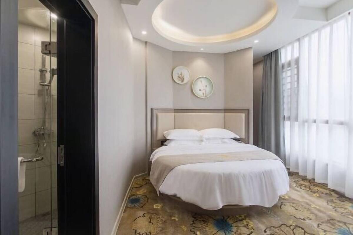 A hotel room with a bed, a shower, a wall clock, and a window with curtains.