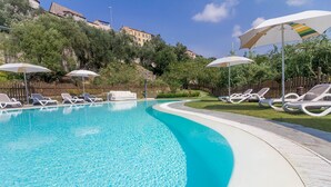 A swimming pool with lounge chairs and umbrellas, surrounded by greenery and buildings in the background. - Housing with private garden terrace and residential swimming pool (Liguria)