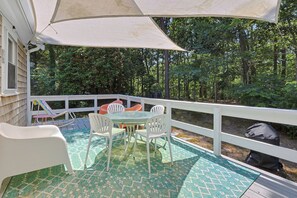 A covered outdoor deck with a table and chairs, surrounded by a white railing, overlooking a wooded area.