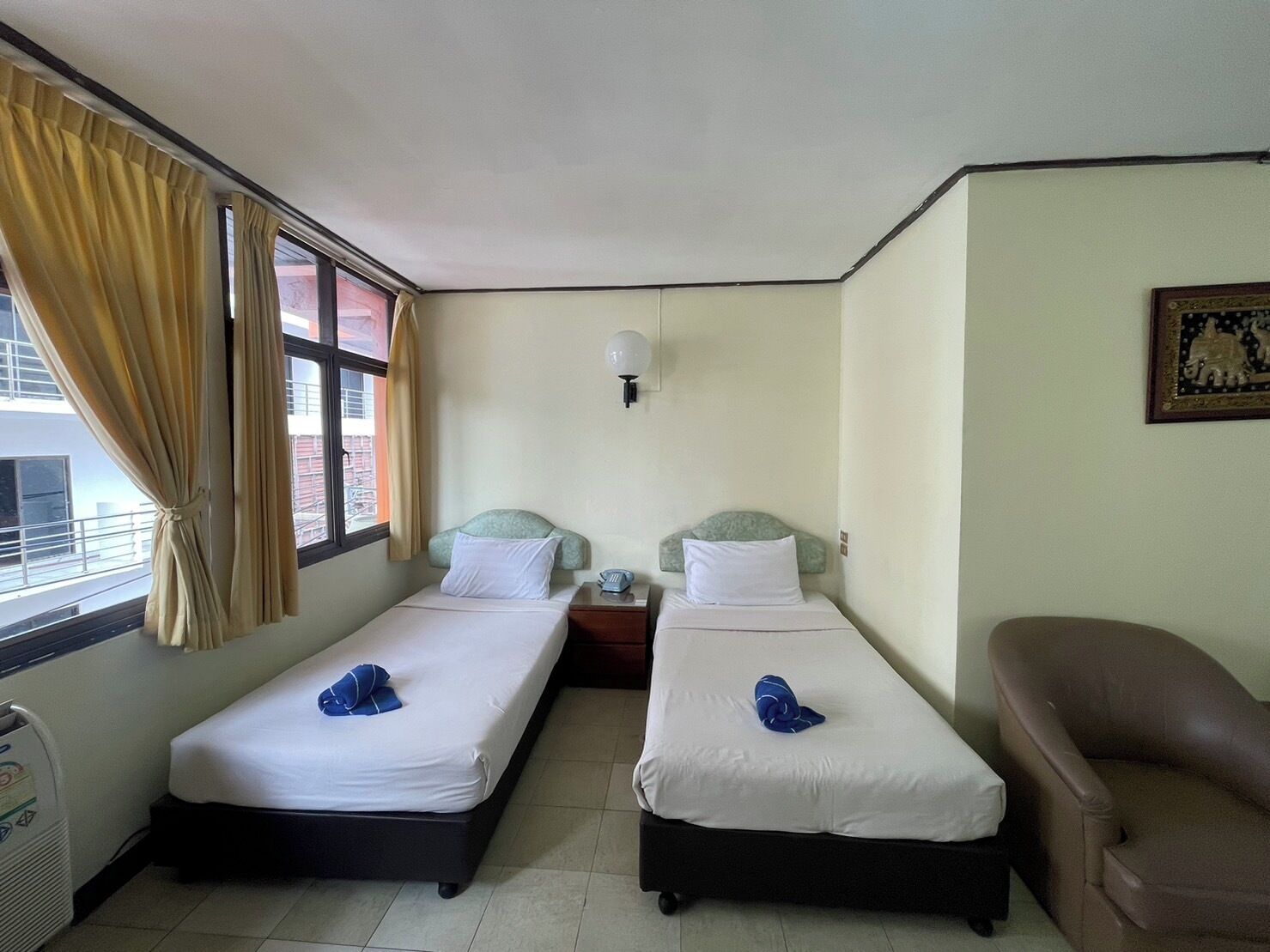 A hotel room with two single beds, a chair, a bedside table, a lamp, and a window with curtains.