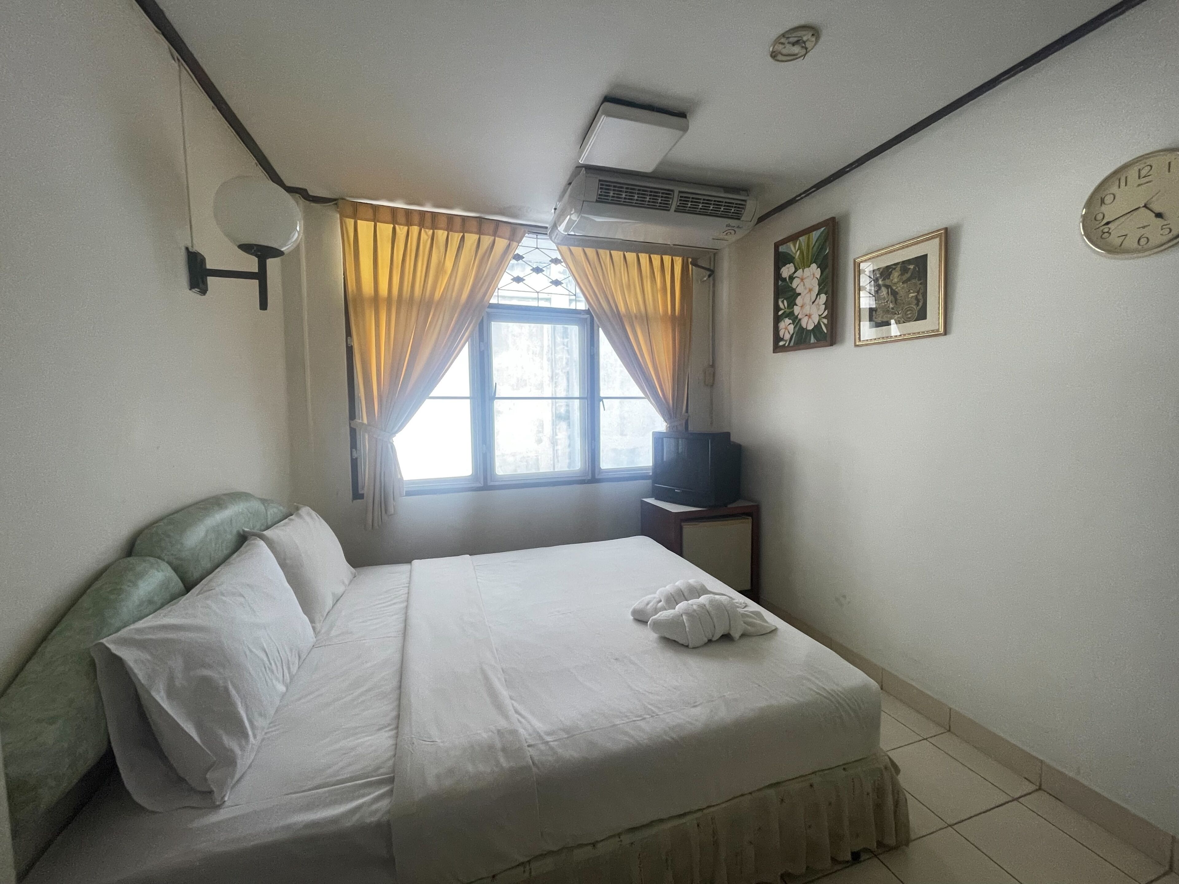A hotel room with a bed, a television on a small table, a window with curtains, and a wall clock.