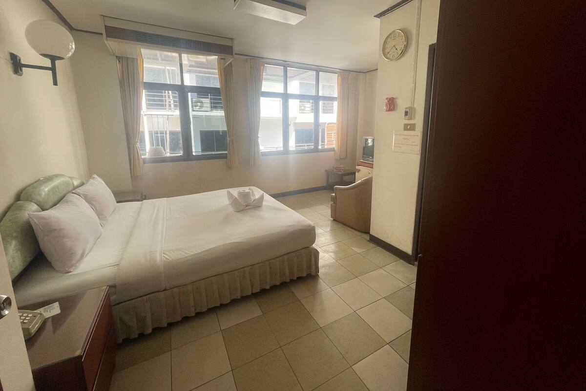 A hotel room with a bed, a small desk with a telephone, a refrigerator, and a window with curtains.