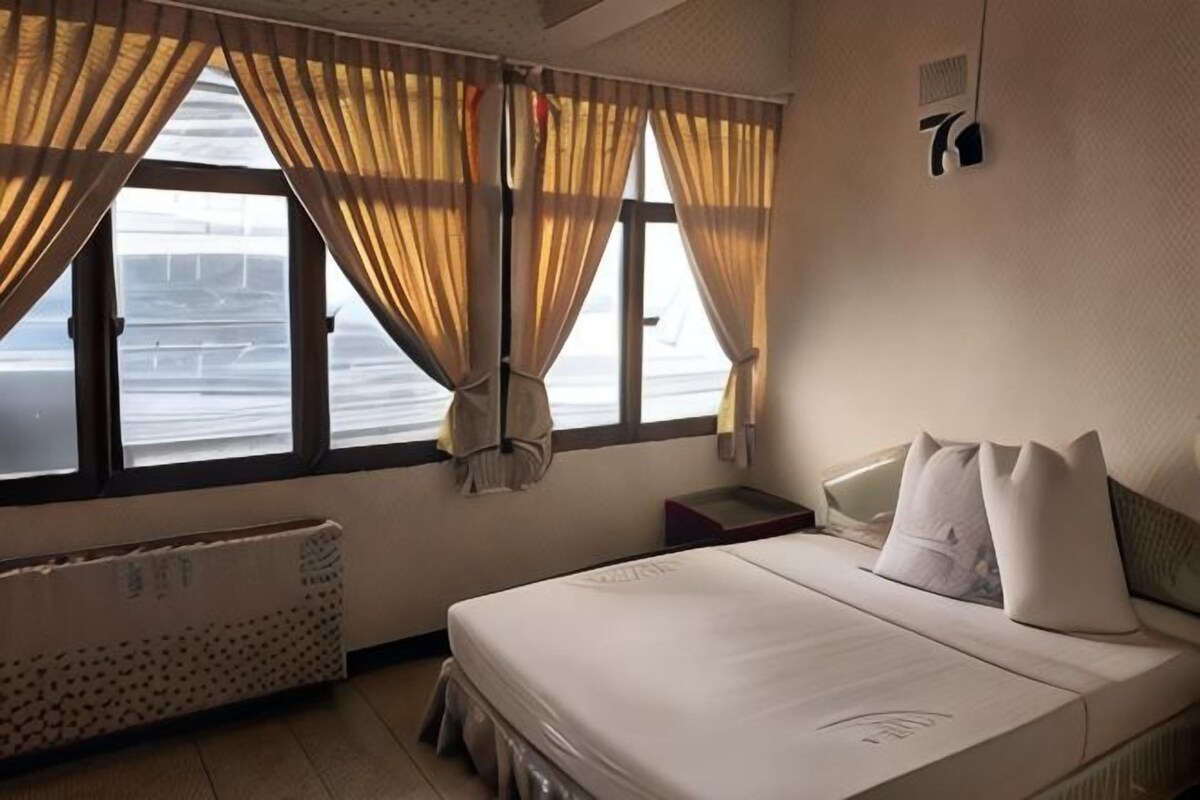 A hotel room with a bed, a window with curtains, a radiator, and a wall-mounted light fixture.
