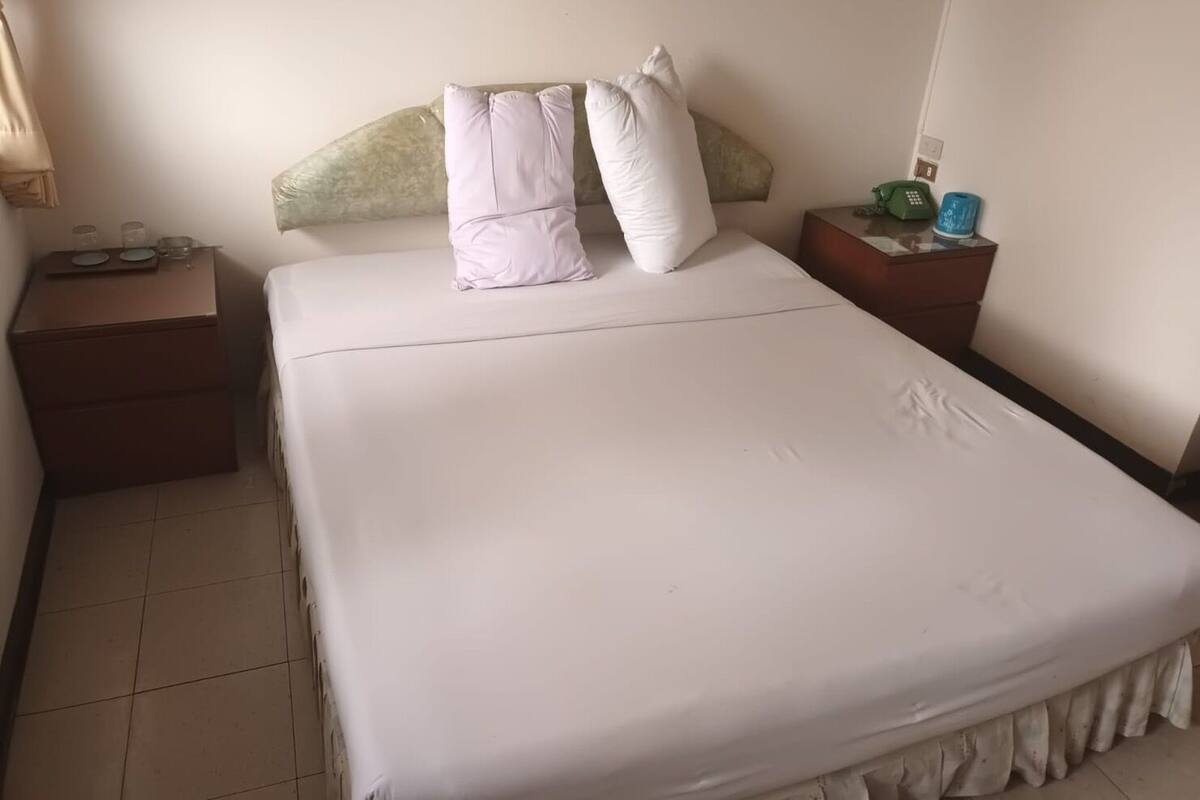 A neatly made bed with white bedding and pillows, a bedside table with a lamp, and a nightstand with a phone and a green bottle.