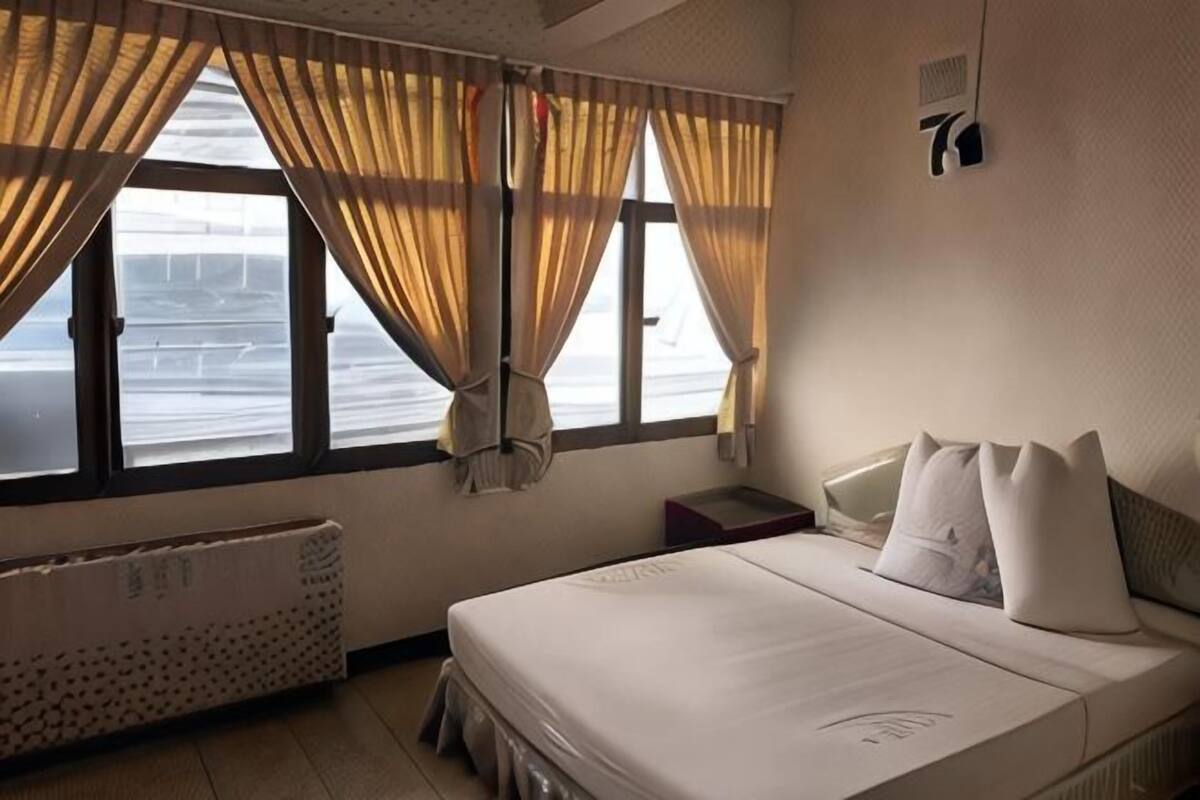 A hotel room with a bed, a window with curtains, a radiator, and a wall-mounted light fixture.