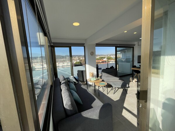 A modern living room with a sofa, a small table, and a view of the cityscape through large windows.