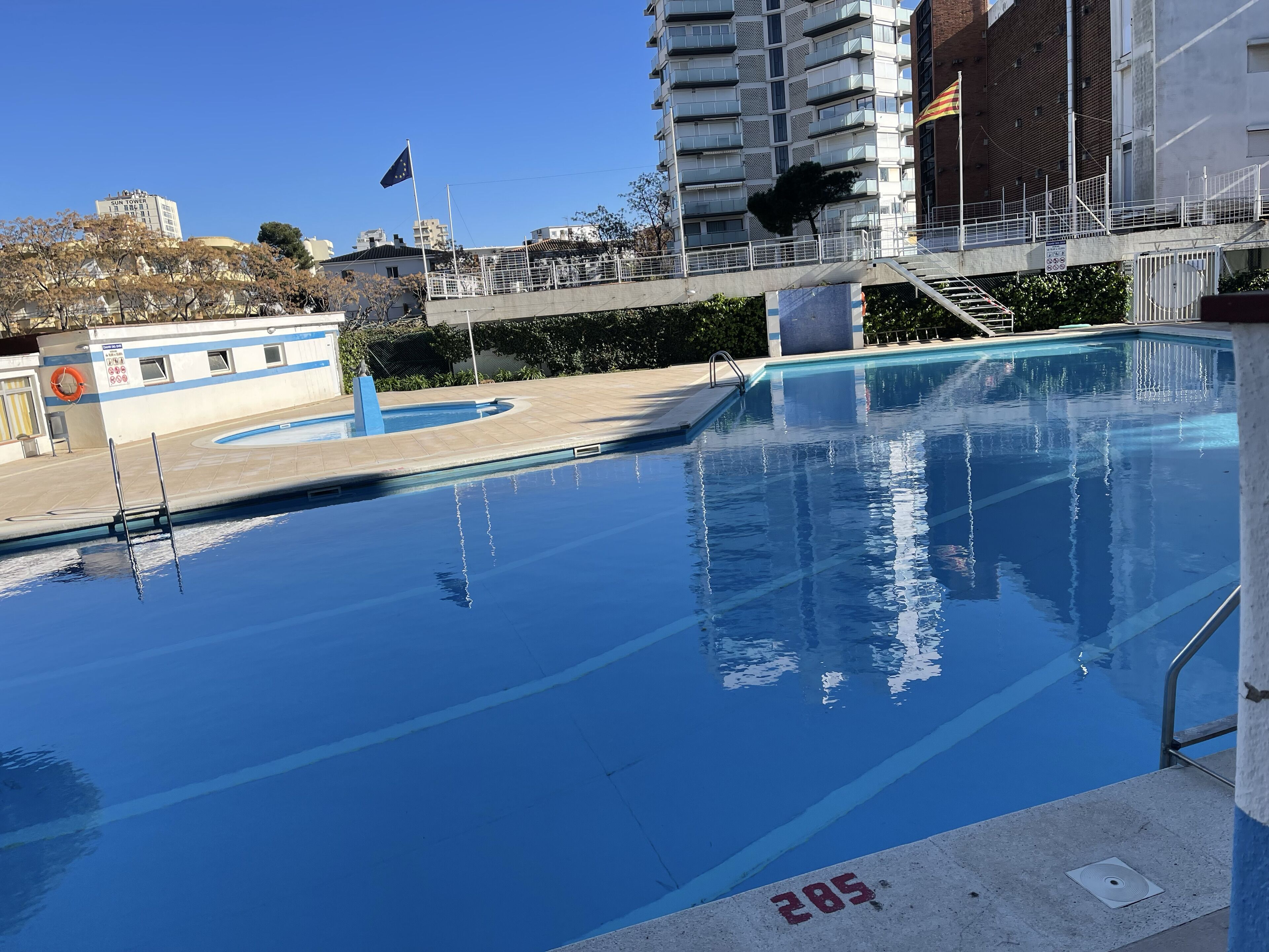 A swimming pool with clear blue water, surrounded by a concrete deck and a metal railing.
