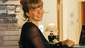Iemand zit aan een bureau met een laptop, een lamp en een plank met diverse spullen op de achtergrond.