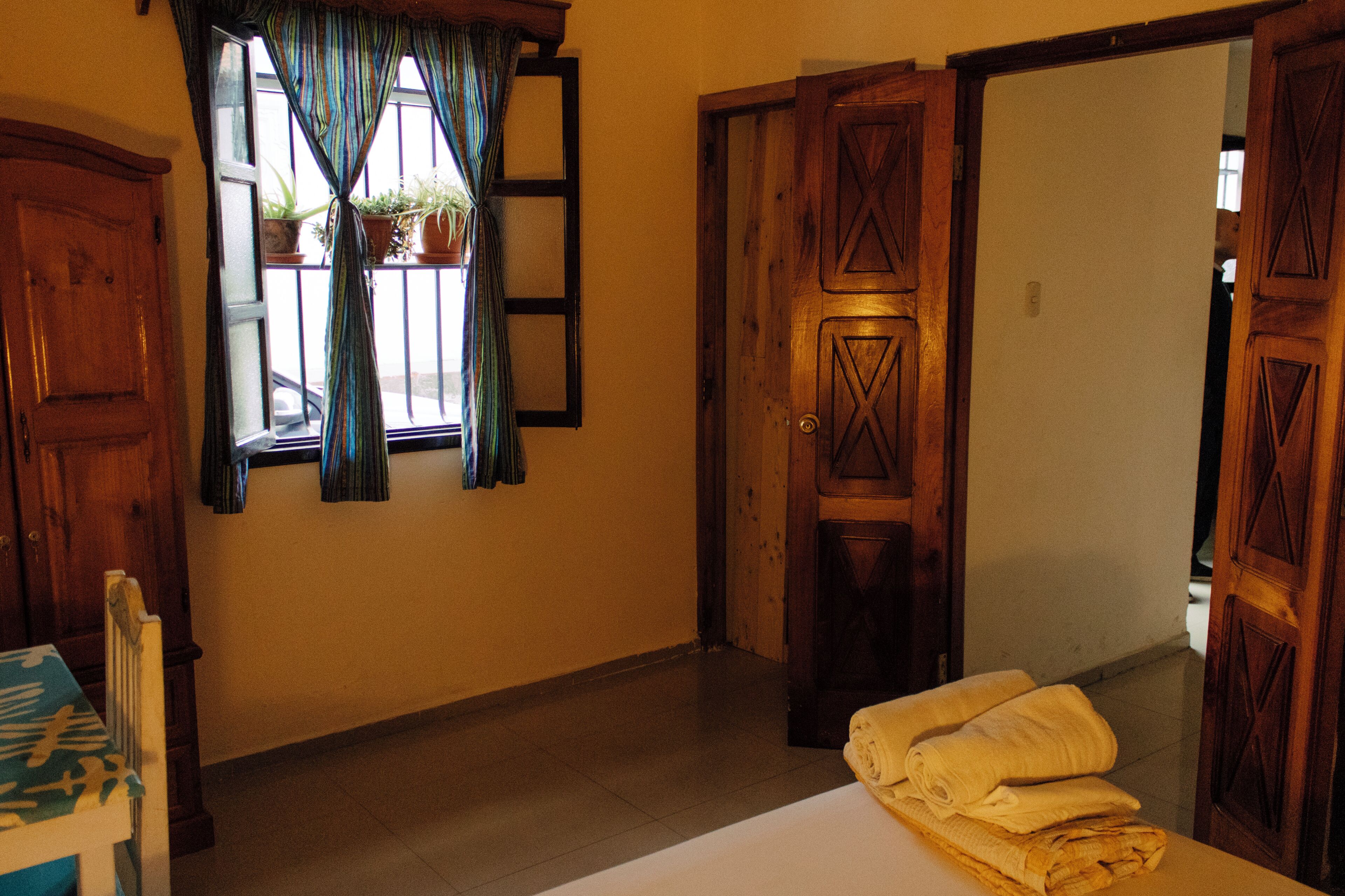 Una habitación con puerta de madera, ventana con cortinas, cama con sábana blanca y una silla con mantel decorado.