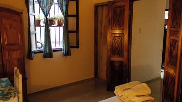 Una habitación con puerta de madera, ventana con cortinas, cama con sábana blanca y una silla con mantel decorado.
