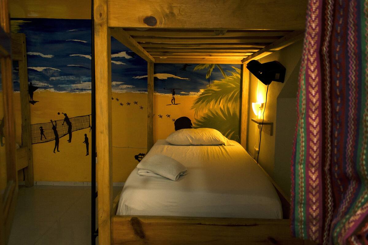 A bunk bed room with a mural of a beach scene, a single bed with a pillow, and a nightstand with a lamp.