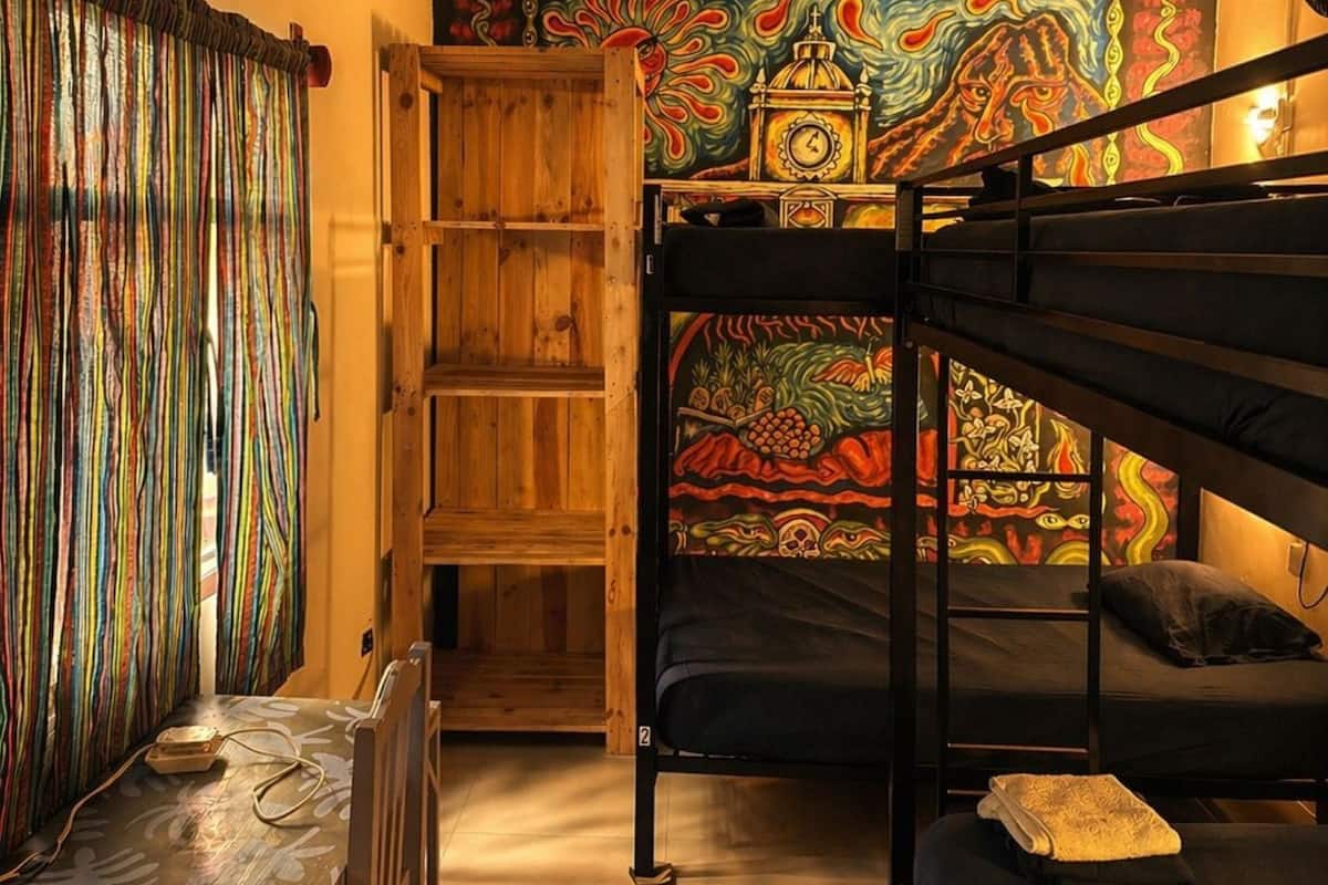 A dormitory room with bunk beds, a desk, a chair, a bookshelf, and a colorful mural on the wall.