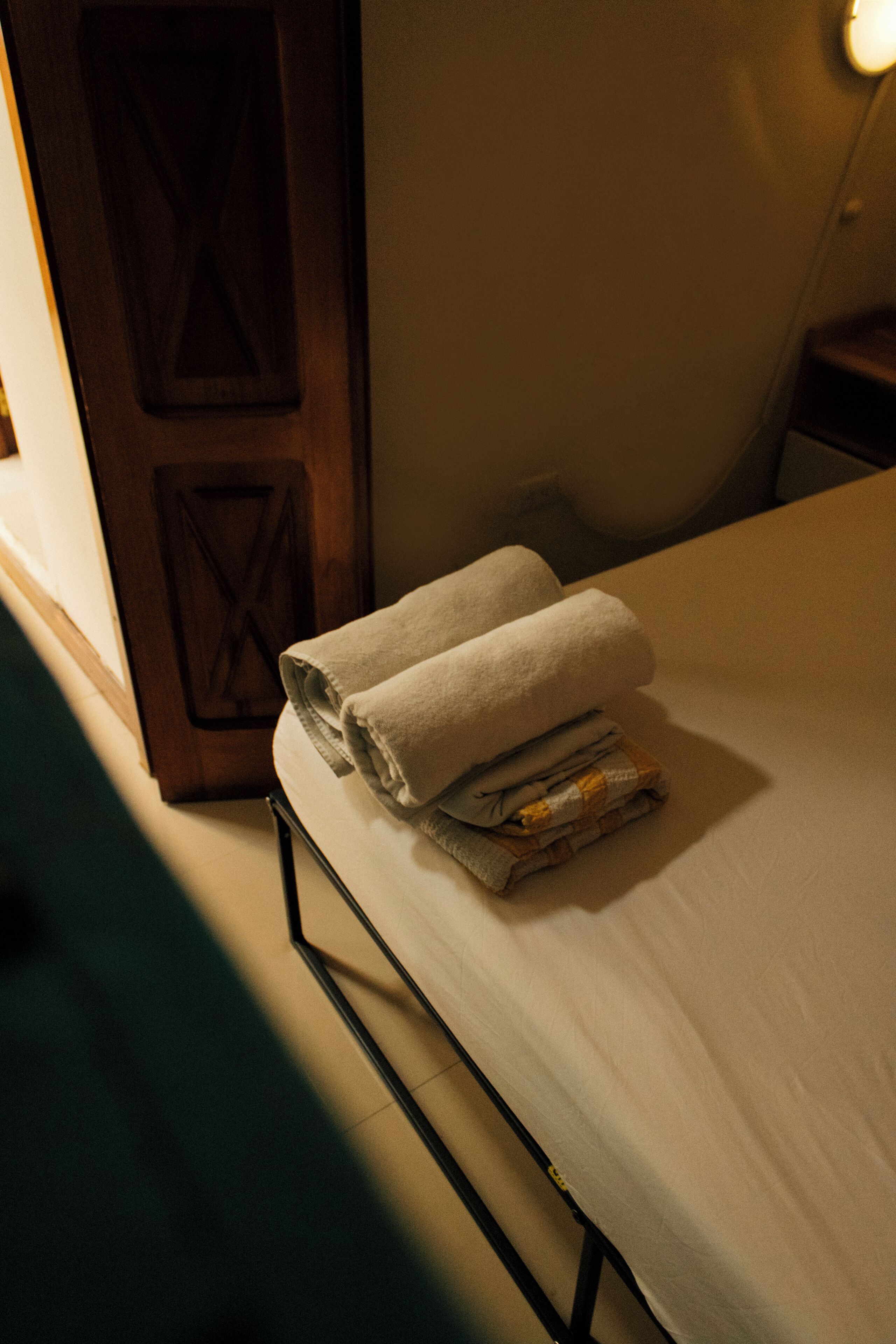 A neatly made bed with folded towels, a wooden door, and a small bedside table.