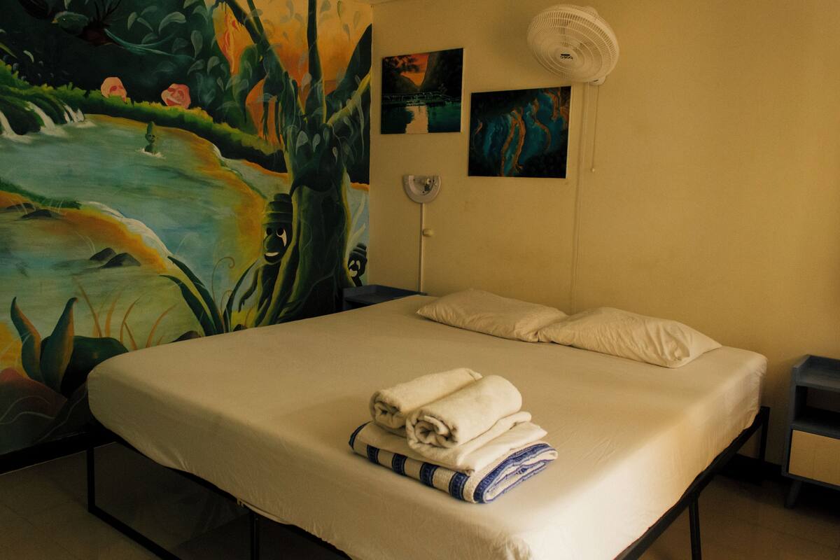 A bed with a white mattress and pillows, a wall with two paintings, a standing fan, and a partial view of a mural.
