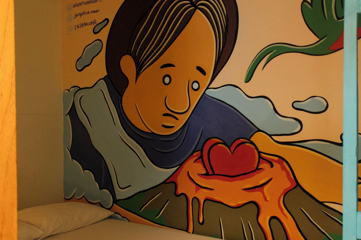 A single bed with a white mattress and a folded towel, a wall with a colorful mural, and a ceiling light fixture.
