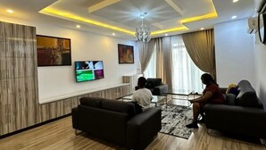 A modern living room with a flat-screen TV, two sofas, a chandelier, and a large window with curtains. - Nigus House Oniru (Lagos)