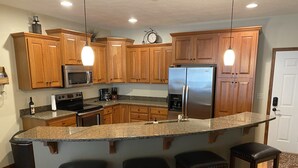 A kitchen with wooden cabinets, a stainless steel refrigerator, a microwave, and a granite countertop. There are three bar stools and a clock on the wall. - Relaxing Getaway, Lakeside Sunsets, 6 Waterpark passes, Memories Galore!! (Arnolds Park)