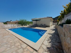 A rectangular swimming pool with a stone border, a covered seating area with chairs, and a stone wall.