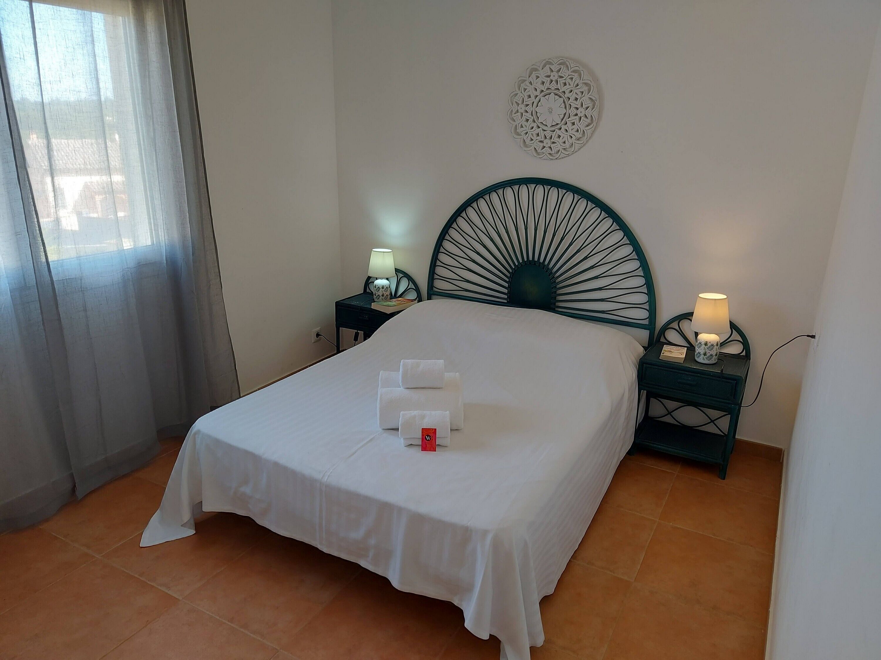 A neatly made bed with a white bedspread and pillows, two bedside tables with lamps, a decorative wall piece, and a window with curtains.