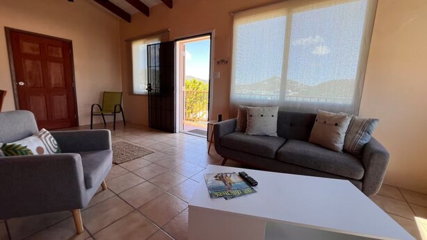 Ein Wohnzimmer mit Sofa, Sessel und Couchtisch. Eine Tür, ein Fenster mit Jalousien und ein Deckenventilator sind ebenfalls vorhanden.